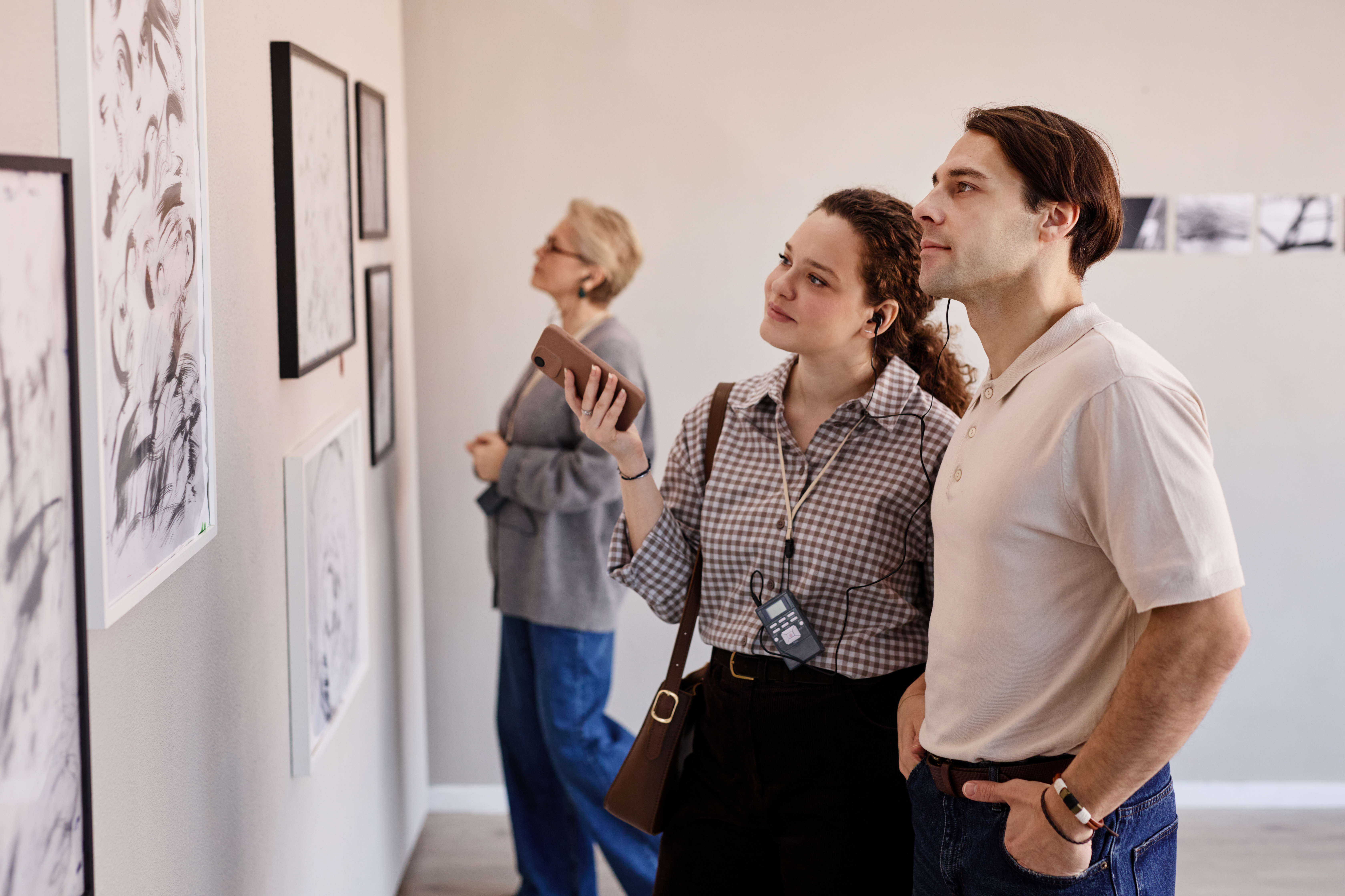 Young Caucasian woman and young Caucasian man standing side by side observing framed artwork on gallery wall, middle aged Caucasian woman in background examining other pieces